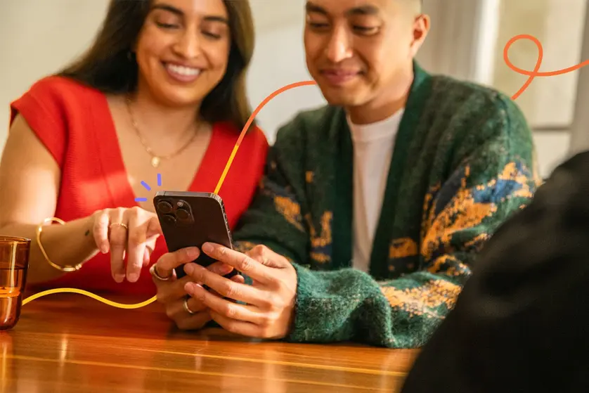An image of a smiling couple sitting at a table looking at a smartphone together.