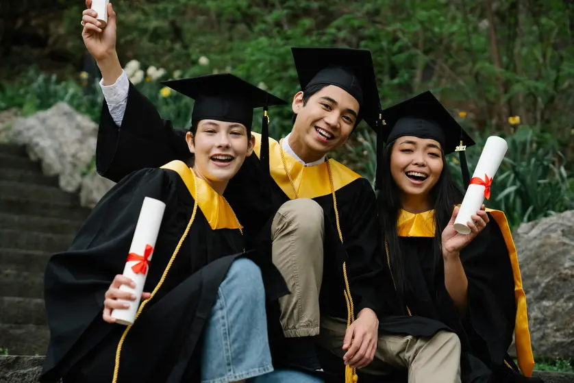 Three college graduates in caps and gowns sitting on outdoor steps, smiling and holding diplomas after their graduation ceremony