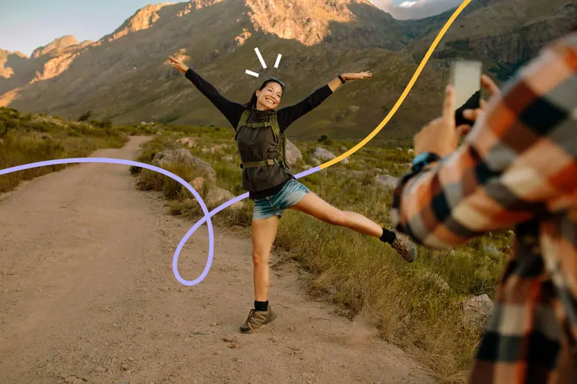 An image of a hiker posing on a mountain trail while a friend takes a photo.