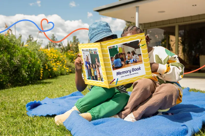 An image of two kids sitting on a blanket outside, reading a yellow memory book.
