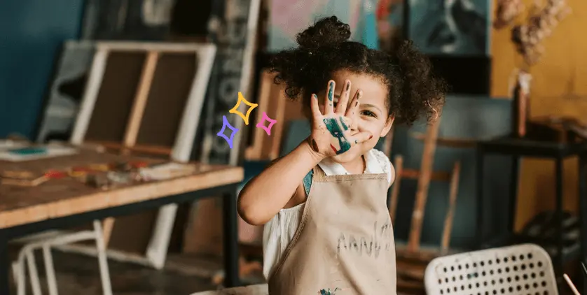 Child smiling with paint-covered hands in an art studio, representing preserving kids’ artwork in a custom photo book.