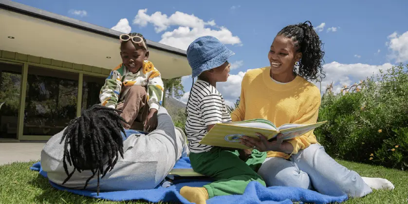 A family of four sits on a picnic blanket in a sunny backyard while an adult reads a picture book to two young children. Everyone is smiling and engaged as they spend time together outside looking through a photo book.